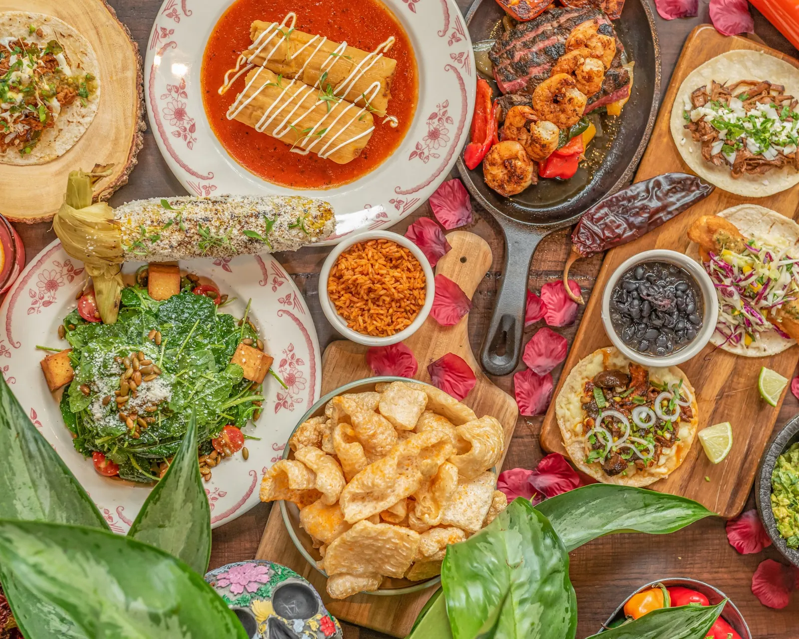 A spread of Mexican dishes on a wooden table: fajitas, tamales, street tacos, grilled corn, beans, rice and a salad with pink flower petals